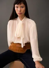 Blusa de viscosa color crema, con diseño semitransparente, cuello con lazada, mangas largas abullonadas y cierre frontal con botones.