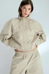 Campera corta de sarga de algodón, color beige, con cuello elevado ajustable por cordón, solapa rompevientos con botones a presión ocultos y cierre. Presenta bolsillos delanteros insertados, mangas ranglán con hombros caídos y bajo globo que genera una silueta abullonada.