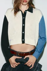 Chaqueta de punto de manga larga con cuello solapa y cierre frontal de botones. Presenta un diseño de bloques de color: cuerpo principal color crema, manga izquierda negra, manga derecha azul claro y cuello negro. Incluye detalles de costura expuesta en azul claro en el cuello y el bajo.