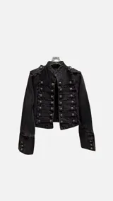 Chaqueta corta de denim negro con diseño de inspiración militar, cuello mao y cierre frontal con múltiples botones metálicos y cordones decorativos. Presenta hombreras y puños con botones a juego.