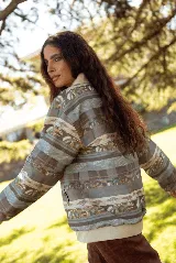 Campera de jean con cierre frontal y cuello tipo bomber, presenta un estampado horizontal con motivos paisajísticos y geométricos en tonos azules, marrones y beige. Posee puños de tela elástica en color blanco.