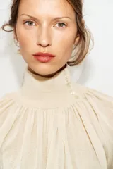 Blusa marrón de punto fluido, con cuello subido abotonado en un lateral y mangas largas con puños ajustados. Presenta un diseño fruncido en la parte frontal que aporta volumen.