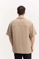 Camisa de manga corta color beige confeccionada en muselina de algodón, con cuello abierto, cierre frontal de botones y un bolsillo aplicado en el pecho.