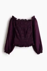 Blusa de color violeta con diseño de hombros descubiertos, cuerpo fruncido y mangas largas abullonadas con puños elásticos. Presenta detalles de volados en el escote, el bajo y los puños.