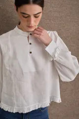 Blusa de lino color blanco con cuello mao, cierre frontal con botones, mangas largas abullonadas con puños ajustados y detalle de volado en el ruedo.