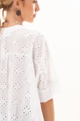 Camisa de broderie blanca con silueta oversize, escote en V, cierre frontal con botones y mangas 3/4. Presenta un diseño con bajo asimétrico, siendo ligeramente más larga en la parte posterior.