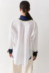 Camisa blanca de corte holgado con cuello y puños tejidos en crochet azul con borde verde. Presenta parches bordados decorativos en el frente y las mangas, cierre frontal con botones y un bolsillo de parche en el pecho.