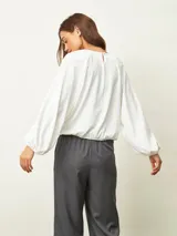 Blusa de color blanco hueso con cuello redondo, mangas largas abullonadas y puños elásticos. Presenta un diseño con frunces en los hombros y ruedo elástico que le otorga una caída holgada.