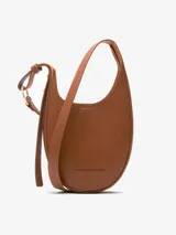 Bandolera pequeña de diseño semicircular con textura granulada, confeccionada en material sintético. Cuenta con un compartimento principal con cierre magnético y asa larga regulable con hebilla metálica. Incluye el logo de la marca grabado en la parte inferior frontal.