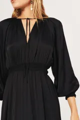 Vestido midi de satén color negro, con escote en V profundo con lazo de ajuste, mangas largas abullonadas con puños elásticos y cintura elástica fruncida.