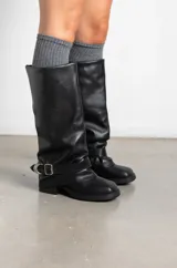 Botas de caña media en cuero negro con diseño arrugado, punta redondeada y suela plana. Presentan una tira decorativa con hebilla metálica y aros en la zona del tobillo.