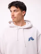 Canguro con capucha y cordones ajustables, de color gris claro. Presenta un logo bordado en el pecho y un parche decorativo en el puño de la manga derecha. Cuenta con un bolsillo frontal tipo canguro y puños acanalados.