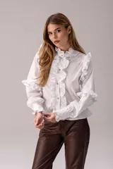 Camisa negra de corte entallado con volados decorativos en el cuello, la botonadura frontal, los hombros y las mangas. Presenta mangas largas con puños ajustados y un ruedo con volado inferior.