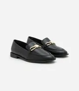 Zapato mocasín femenino de PU color negro, con punta cuadrada y detalle metálico dorado en el empeine. Tiene un tacón bajo y suela de goma.