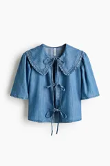 Blusa corta de denim liviano con mangas cortas abullonadas, cuello Peter Pan con volados y cierre frontal con tres tiras para atar.