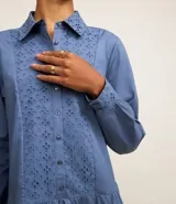 Vestido corto de corte evasé confeccionado en tricolina de algodón, color azul. Presenta cuello camisero, cierre frontal de botones y un panel central con detalle bordado calado. Posee mangas largas con puños abotonados.
