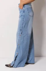Pantalón de jean estilo wide leg color celeste claro, con tiro alto y cierre frontal con botón. Presenta una abertura lateral en cada pierna decorada con botones metálicos a lo largo de toda la extensión.