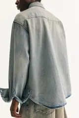 Camisa vaquera de algodón rígido, color celeste lavado, con cuello, botones a presión frontales y en los puños, canesú delantero y trasero, y dos bolsillos superiores con solapa y botón a presión. Presenta un bajo redondeado y un corte oversized.