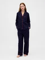 Conjunto de pijama compuesto por una camisa de manga larga con cuello solapa, cierre frontal de botones y bolsillo en el pecho, y un pantalón de corte recto con cintura elástica. Ambas piezas presentan un estampado de pequeños motivos oscuros sobre fondo claro y detalles de ribetes en contraste.