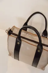 Cartera tipo bowling color beige con detalles en cuero negro, doble asa corta y cierre superior con cremallera.