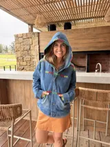 Campera de jean azul con capucha y detalles de bordado artesanal multicolor en la capucha, el pecho, los bolsillos y las mangas. Cuenta con cierre frontal de botones, bolsillos delanteros y diseño cómodo.
