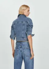 Chaqueta vaquera de corte corto y diseño recto, confeccionada en tejido de algodón con elastano. Presenta cuello de solapa, cierre frontal de botones metálicos, bolsillos con solapa en el pecho y bolsillos laterales. Las mangas largas cuentan con puños abotonados.