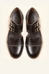 Zapato tipo oxford abierto en los laterales, confeccionado en cuero vacuno negro. Presenta doble tira con hebillas metálicas en el empeine y detalle de tachas plateadas en el contorno del talón y la puntera.