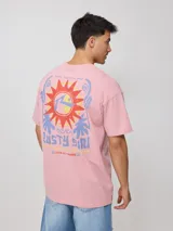 Remera de algodón blanca de corte oversize, con mangas cortas y cuello redondo. Presenta un gran estampado multicolor en la espalda con diseño gráfico de sol y texto, y un estampado más pequeño en el frente.