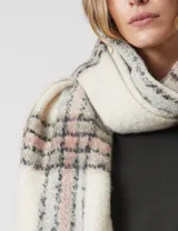 Bufandón de tejido soft, color crema con patrón a cuadros en tonos rosa pálido, gris y negro. Presenta terminación con flecos.