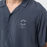 Camisa de manga corta en color gris oscuro, confeccionada en viscosa con cuello tipo cubano y cierre frontal de botones. Presenta un pequeño estampado circular en el pecho con la inscripción Black.