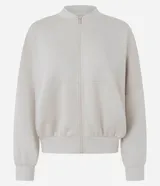 Campera bomber deportiva de mujer color beige, confeccionada en modal con cierre frontal por cremallera, cuello medio y bolsillos. Presenta mangas largas con puños elásticos y elástico en la cintura.