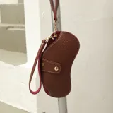 Estuche para lentes de diseño ergonómico, confeccionado en ecocuero texturizado color marrón con interior en contraste color fucsia. Cuenta con cierre mediante broche a presión metálico y correa de mano extraíble con herrajes dorados.