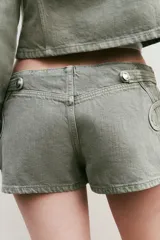 Short mini de tiro bajo color verde oliva, con detalle de botones metálicos y aplicaciones decorativas en el mismo tejido. Cuenta con cierre frontal oculto.