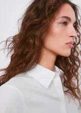 Camisa de popelín blanco entallada, con cuello camisero, mangas largas y cierre frontal de botones ocultos.