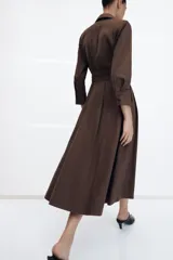 Vestido midi de estilo camisero color verde oscuro, con cuello solapa, escote en pico y cierre frontal con botones. Presenta manga larga, bolsillos laterales ocultos, solapas decorativas en el pecho y cinturón de lazada en el mismo tejido.