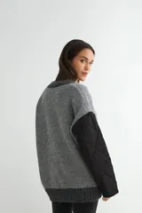 Saco tipo cardigan de punto gris con diseño híbrido. Presenta mangas largas y bolsillos frontales de tela técnica acolchada en color negro, con cierre frontal de botones y bordes acanalados.