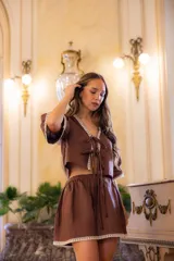 Top de lino color marrón con escote en V, mangas tres cuartos y cierre frontal mediante lazos. Presenta detalles de puntilla cruda en el escote, puños y ruedo.