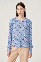 Blusa celeste con estampado de flores rosas y negras, de mangas largas abullonadas y cuello redondo con abertura en forma de gota.