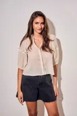 Blusa de color arena con cuello en V, mangas tres cuartos abullonadas y detalles de encaje bordado en hombros y mangas. Presenta un diseño ligero y delicado con cierre frontal.