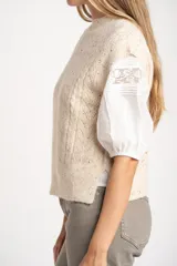 Chaleco de tejido de punto color beige melange con textura tweed y cuello redondo. Presenta un patrón de rombos calados y trenzas.