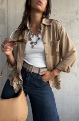 Sobrecamisa de gamuza color beige, con cuello camisero, cierre frontal con botones metálicos y bolsillo en el pecho. Presenta un diseño con calados florales y bordes ondulados en la parte inferior.