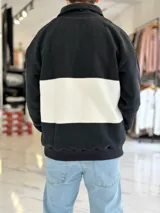 Buzo de algodón negro con una franja horizontal blanca en el centro, cuello solapa con medio cierre y puños y cintura elásticos acanalados. Cuenta con pequeños estampados en el pecho.