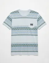 Remera de manga corta con estampado geométrico de líneas y triángulos en tonos celestes y verdes sobre fondo gris claro. Presenta un bolsillo frontal en el pecho con una etiqueta de marca.