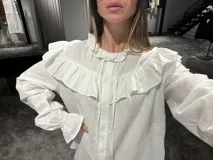 Camisa blanca de textura rugosa con volados en el pecho y hombros, cuello redondo con lazos de ajuste, cierre frontal con botones y mangas largas con puños con volados.