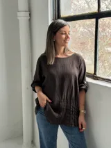 Blusa de lino color marrón chocolate, de corte holgado, con cuello redondo, mangas tres cuartos con trabilla y botón para sujetar remangadas, y detalle de alforzas verticales en el frente.
