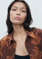 Camisa marrón de algodón con estampado de palmeras, cuello camisero, manga larga y cierre frontal de botones.
