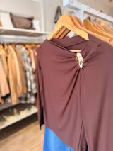 Remera de manga larga color chocolate, con escote drapeado asimétrico y un aplique metálico decorativo en el hombro.