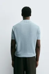 Polo de manga corta, color celeste pálido, con cuello solapa y abertura frontal en V. Tejido de punto con textura acanalada vertical.