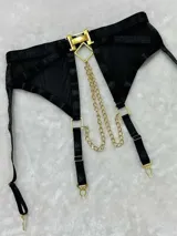 Conjunto de lencería de cuatro piezas en color negro, confeccionado con bandas elásticas y malla transparente. Incluye soutiene con aro, tiras cruzadas decoradas con tachas metálicas, cadenas y un dije romboidal dorado en el centro. La parte inferior presenta bombacha tipo tanga con tiras ajustables, arnés de piernas y portaligas incorporado con herrajes dorados.