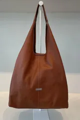 Bolso tipo tote de gran capacidad, confeccionado en cuero vacuno color suela. Presenta un diseño minimalista con costura central vertical, asas integradas y placa metálica con logo de la marca en la parte frontal. Incluye fuelles laterales ajustables y sobre interior desmontable.